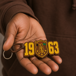 Load image into Gallery viewer, Hand holding a keychain with '1963' and a logo, against a brown background.
